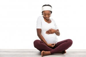 Woman wearing a white headband and a white shirt is pregnant and sitting on the floor while smiling and holding her stomach.