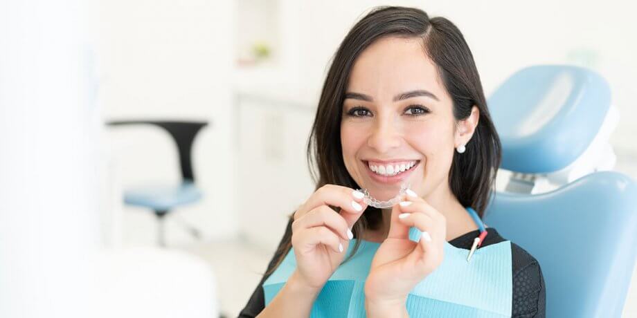 a woman holds a clear aligner and smiles from the dentist chair