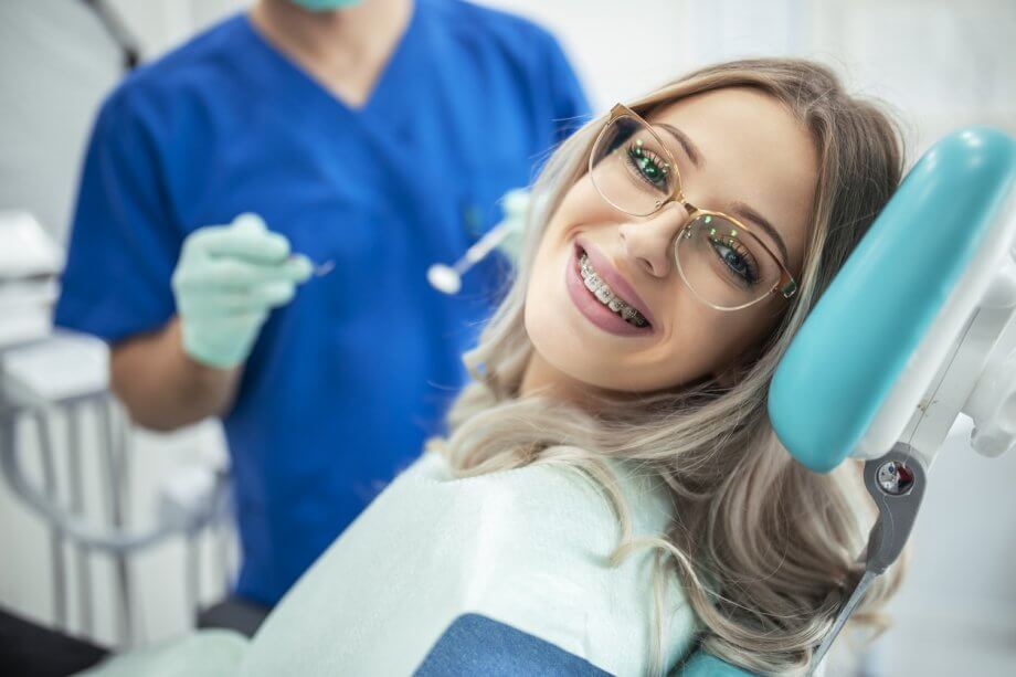 Young girl with long wavy blonde hair, glasses, & braces smiling while in a dentist chair as a hygienist stands behind her.
